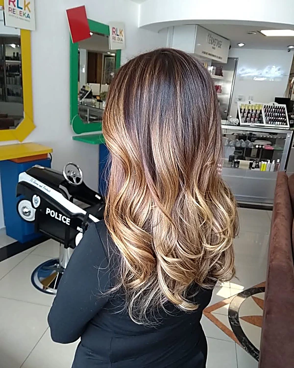 Balayage dorado en ondas largas, de espaldas; al fondo, la silla de patrulla del salón